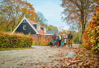 Gezin wandelt met hond bij vakantiehuisje in een herfstig vakantiepark, omringd door kleurrijke bomen.