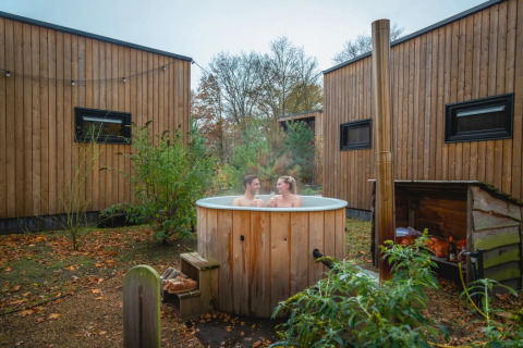 Twee mensen ontspannen in een houten hot tub bij glamping huisjes in een natuurvriendelijk vakantiepark.