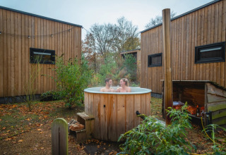 Twee mensen ontspannen in een houten hot tub bij glamping huisjes in een natuurvriendelijk vakantiepark.