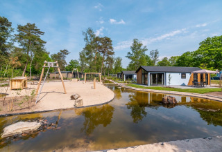 Zicht op een glamping vakantiepark met moderne huisjes, speeltuin en vijver omringd door bomen onder blauwe lucht.