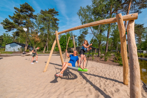 Kinderen en een vrouw spelen op een schommel in een vakantiepark met glamping, zand en bomen.