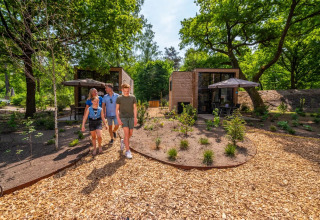Groep mensen wandelt over een houtpad langs moderne glampinghuisjes in een groen vakantiepark.