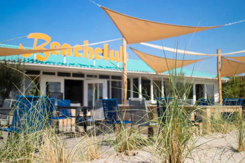 Een zonnige glamping beachclub bij een vakantiepark, met zand, winddoeken en blauwe stoelen buiten.