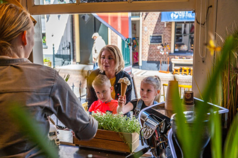 Twee kinderen en een vrouw kopen ijsjes bij een raam in een gezellige omgeving van een vakantiepark.