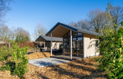 Een moderne lodge met grote ramen, omringd door bomen en struiken onder een heldere blauwe lucht.