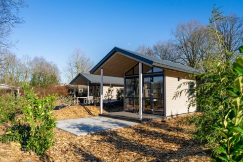 Een moderne lodge met grote ramen omgeven door bomen en struiken onder een heldere blauwe lucht.