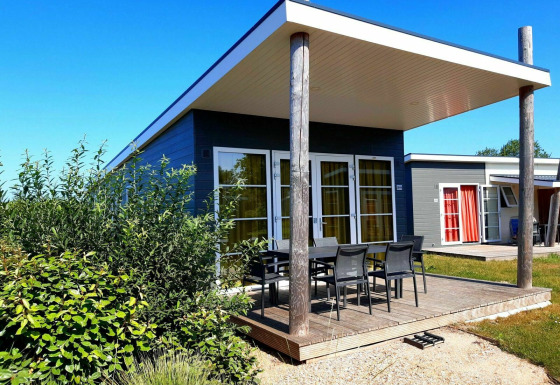 Foto van een moderne lodge met houten veranda, tuinstoelen, veel groen en een stralend blauwe lucht.