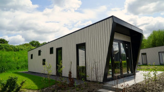 Moderne lodge met schuine gevel en grote ramen, omringd door gras en struiken onder een wolkenlucht.