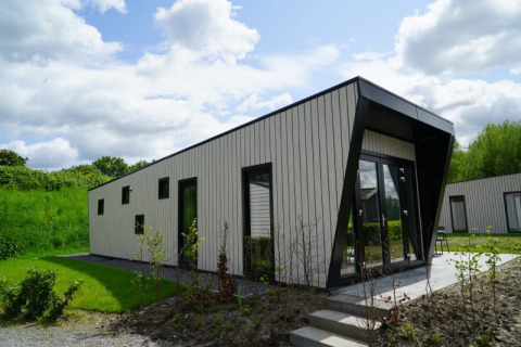 Moderne Lodge mit schrägem Dach und großen Fenstern, umgeben von Grünflächen und bewölktem Himmel.