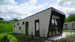 Lodge moderne avec toit incliné et grandes fenêtres, entouré de verdure sous un ciel partiellement nuageux.