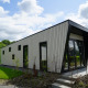 Moderne lodge med skråt tag og store vinduer, omgivet af grønne buske og græs under en skyet himmel.