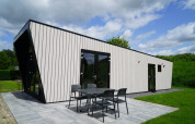 Moderne lodge med sort-hvide vægge, store vinduer og udendørs spiseplads omgivet af græs og natur.