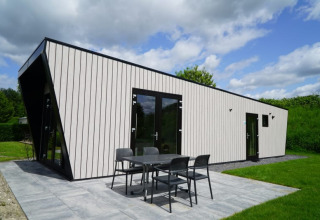 Lodge moderno con rivestimento chiaro, infissi neri, ampie vetrate e tavolo esterno immerso nel verde.