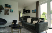 Moderne lodge woonkamer met sofa, bijzettafels, wandkunst, kitchenette en grote ramen buiten.