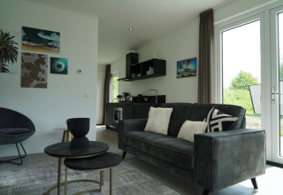 Moderne lodge woonkamer met sofa, bijzettafels, wandkunst, kitchenette en grote ramen buiten.