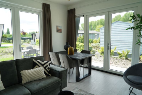 Moderne lodgewoning met grote ramen, eettafel, grijze zetel en zicht op tuin en terras buiten.