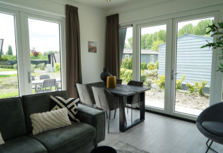 Moderne lodgewoning met grote ramen, eettafel, grijze zetel en zicht op tuin en terras buiten.