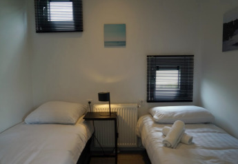 Eenvoudige lodge slaapkamer met twee eenpersoonsbedden, een tafeltje, lamp en zacht daglicht.