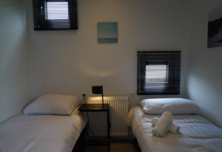 Chambre simple dans un lodge avec deux lits simples, petite table, lampe et lumière douce aux fenêtres.