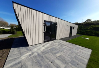 Moderne lodge met schuin dak, grote glazen deuren en een betegeld terras met gras en blauwe lucht.