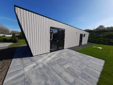 Moderne lodge met schuin dak, grote glazen deuren en een betegeld terras met gras en blauwe lucht.