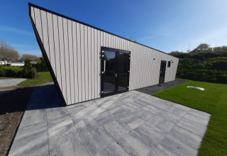 Moderne lodge met schuin dak, grote glazen deuren en een betegeld terras met gras en blauwe lucht.