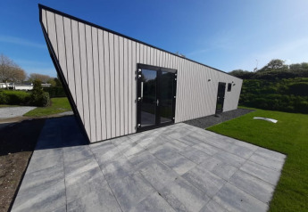 Lodge moderne au toit incliné avec grandes portes vitrées, terrasse carrelée et pelouse verte autour.