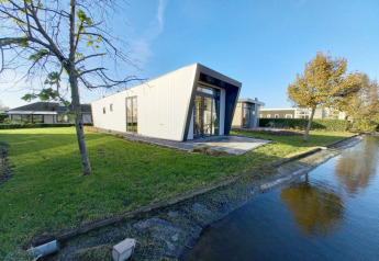 Modern lodge with a tilted facade beside a canal, surrounded by green grass and trees under clear sky.