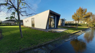 Lodge moderne à façade inclinée près d’un canal, entouré de pelouse verte et d’arbres sous ciel bleu.