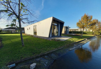 Moderne lodge med vinklet facade ved en kanal, omgivet af grønne træer og græs under blå himmel.