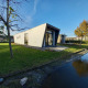 Moderne lodge med vinklet facade ved en kanal, omgivet af grønne træer og græs under blå himmel.