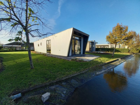 Moderne lodge met schuine gevel aan het water, omgeven door gras en bomen onder een heldere lucht.
