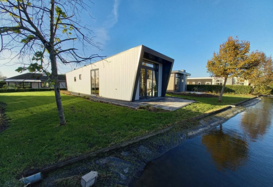 Moderne lodge met schuine gevel aan het water, omgeven door gras en bomen onder een heldere lucht.