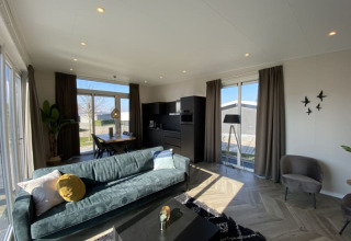 Moderne lodge-living met grote ramen, groene sofa, zwarte keuken, eettafel en veel natuurlijk licht.