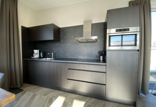 Cocina moderna en una cabaña con muebles oscuros, horno integrado, cafetera y luz natural.