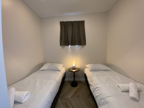 Sencilla habitación de lodge con dos camas individuales, sábanas blancas, mesa central y ventana pequeña.