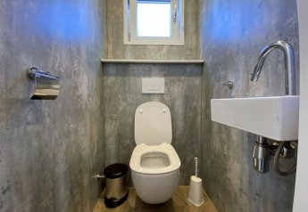 Modern lodge restroom with wall-mounted toilet, compact sink, window, toilet brush, and paper holder.