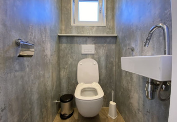 Moderne lodge-toilet med ophængt toilet, lille håndvask, spejl, toiletpapirholder og vindue.