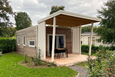 Gezellige houten lodge met overdekt terras, tafel en stoelen, omgeven door een groene tuin en struiken.