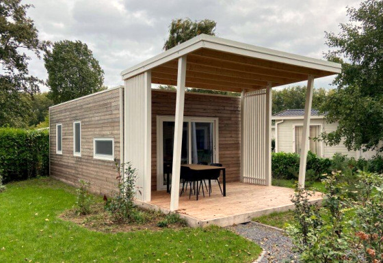 Gezellige houten lodge met overdekt terras, tafel en stoelen, omgeven door een groene tuin en struiken.