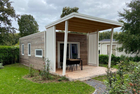Lodge en bois moderne avec terrasse couverte, table, chaises et jardin verdoyant en arrière-plan.