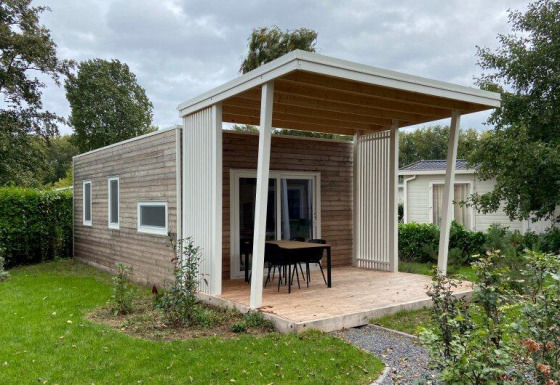 Lodge en bois moderne avec terrasse couverte, table, chaises et jardin verdoyant en arrière-plan.