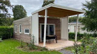 Acogedora cabaña de madera con porche cubierto, mesa exterior y sillas. Jardín verde alrededor.