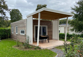 Et hyggeligt træhus med overdækket terrasse, bord og stole, omgivet af frodige grønne planter og hæk.