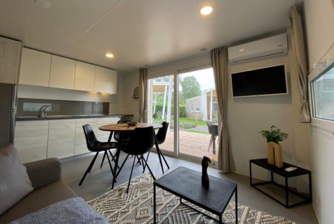 Moderne glamping-opholdsstue med køkken, spiseplads og udsigt over udendørs terrasse gennem glasdøre.