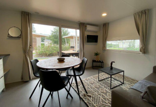 Lyst glampingindkvartering med spisebord, sorte stole, sofabord og store vinduer med udsigt til naturen.