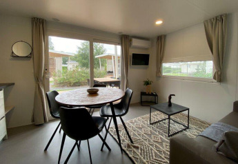 Lyst glampingindkvartering med spisebord, sorte stole, sofabord og store vinduer med udsigt til naturen.