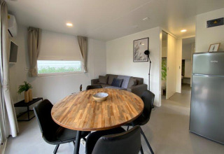 Modern glamping accommodation living area with wood dining table, sofa, fridge, and bedroom entrance.