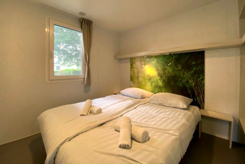 Knusse glampingkamer met tweepersoonsbed, witte lakens, opgerolde handdoeken en natuurhoofdboard.