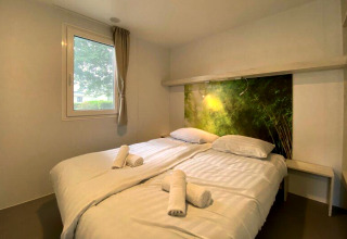 Knusse glampingkamer met tweepersoonsbed, witte lakens, opgerolde handdoeken en natuurhoofdboard.